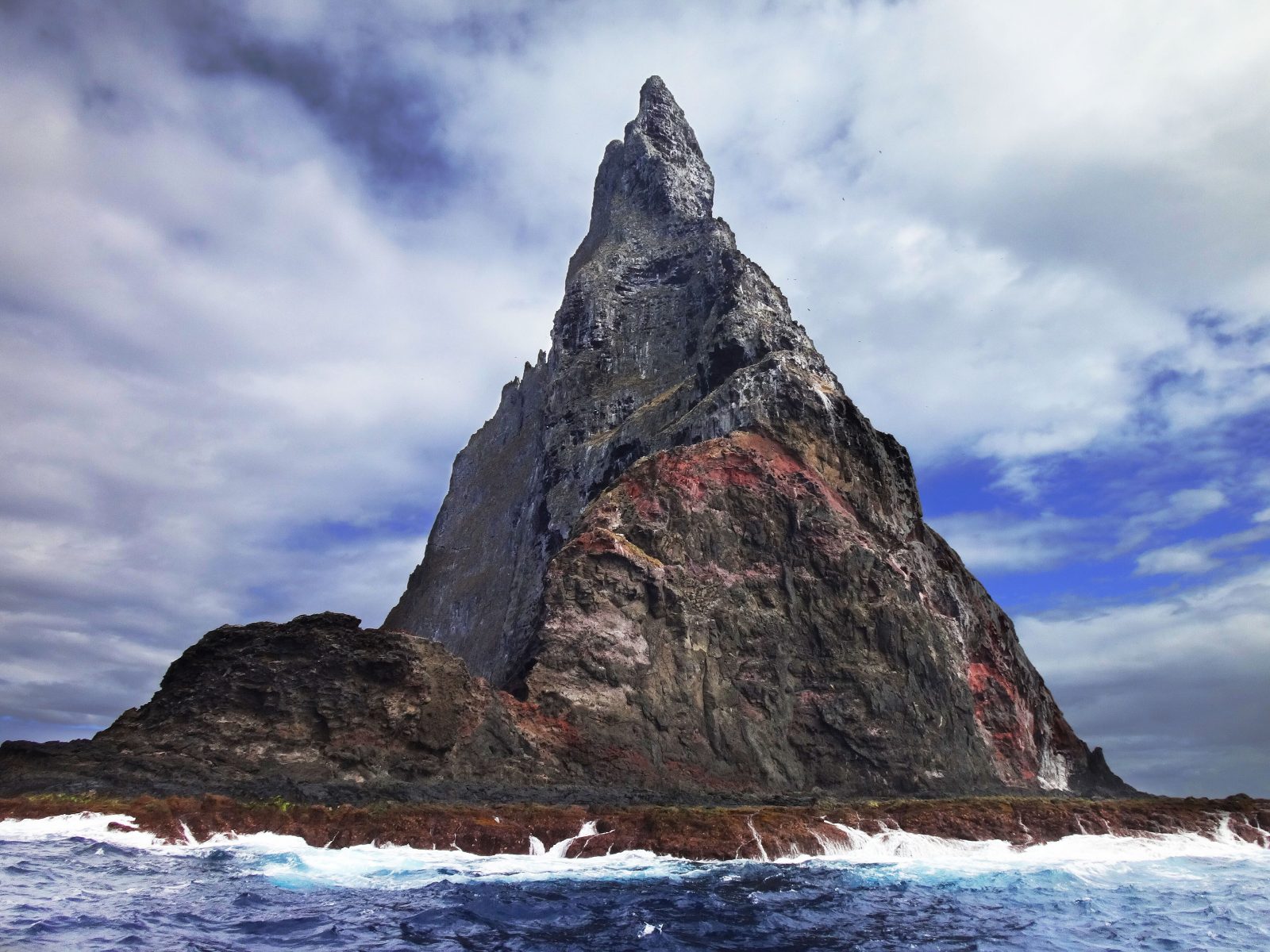 Smailiaviršūnė tamsi uola rausvais šlaitais kyla iš banguojančio vandenyno po debesuotu dangumi