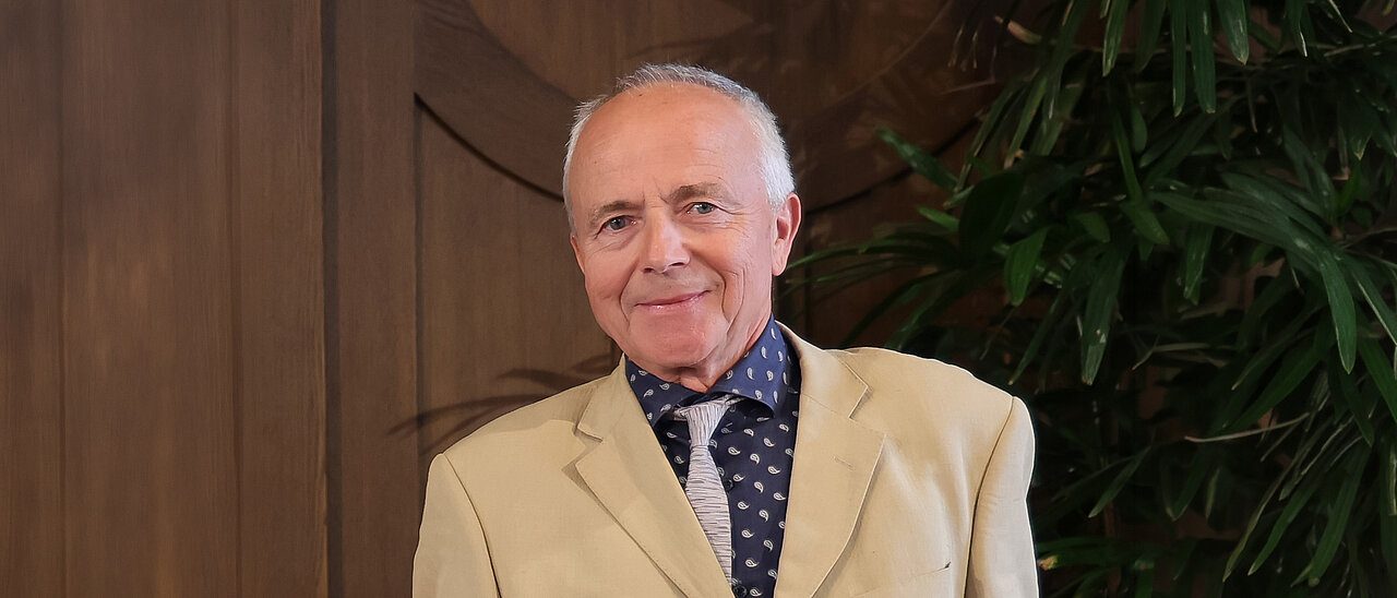Georgas Nagelis šviesiu švarku šypsosi prie medinės sienos