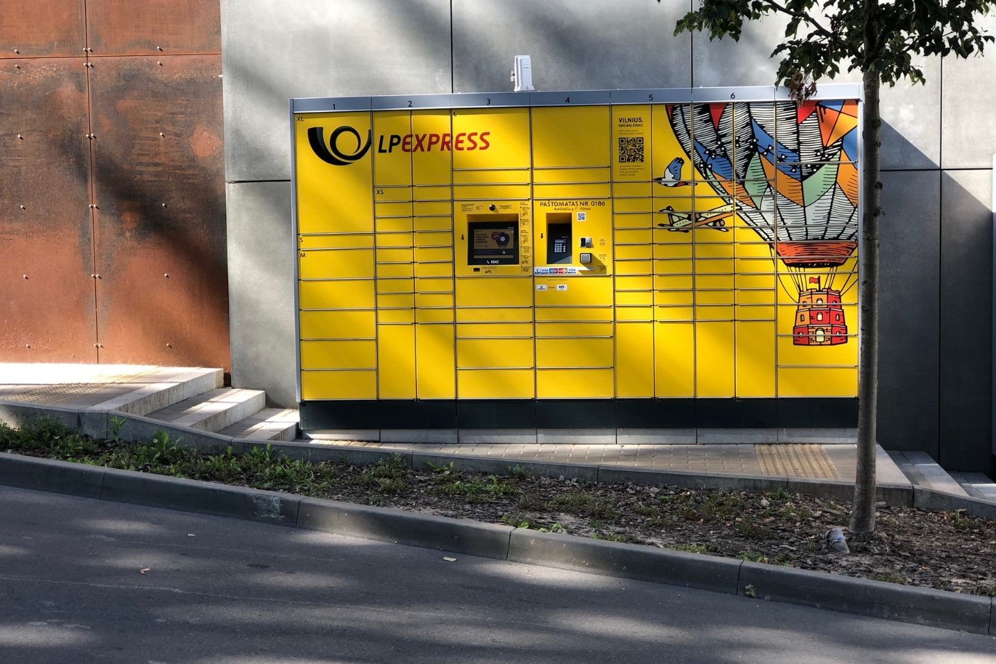 Geltonas LP EXPRESS paštomatas su spalvingu oro baliono piešiniu prie pastato sienos, priekyje šaligatvis