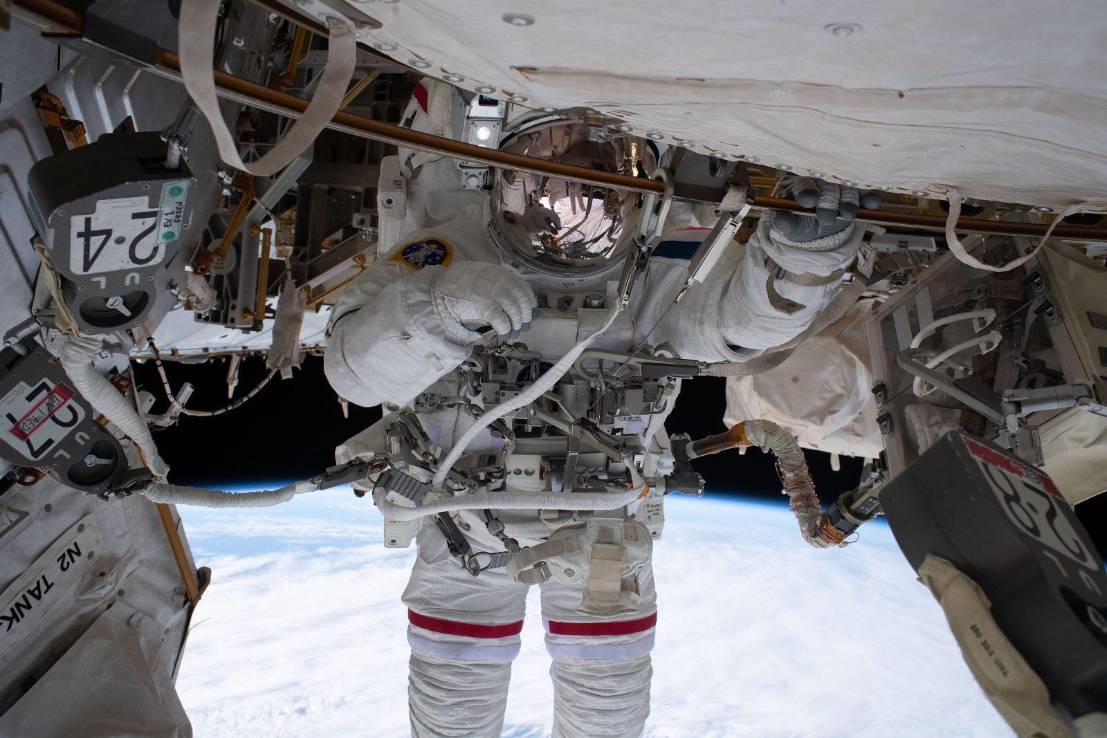 Astronautė baltu skafandru su nuleistu auksiniu šalmo skydeliu dirba prie kosminės stoties, apačioje matyti Žemė