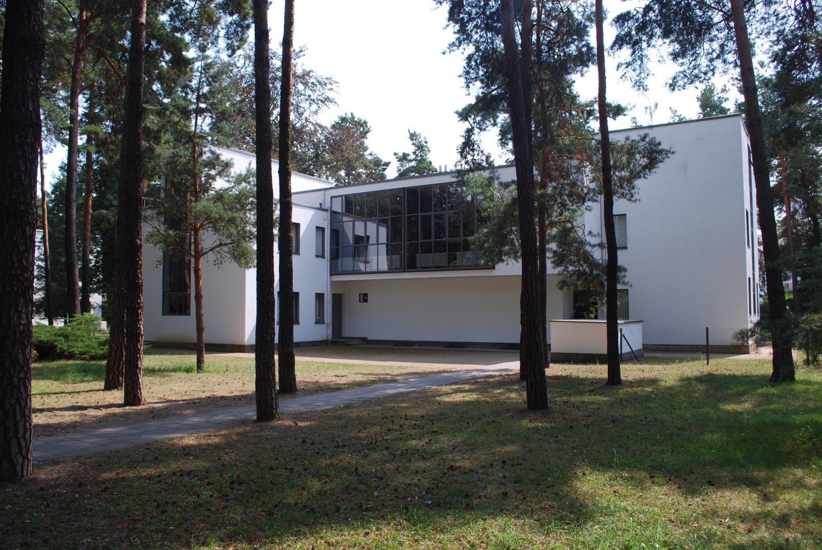 Baltas kubinis dviejų aukštų namas su stiklinta laiptine, apsuptas pušų ir vejos